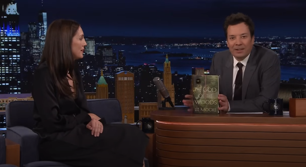 image of Liz Moore sitting in a blue dress looking at Jimmy Kimmel as he holds her book up towards the camera